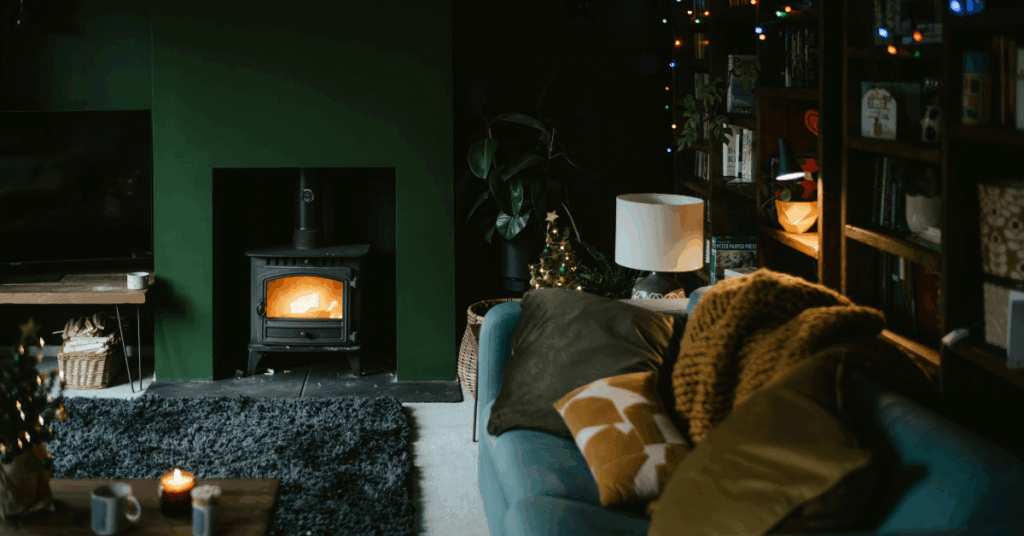 a cosy living room with a green fire surround and log fire, sofa full of comfy cushions and throw and fairy lights hanging above
